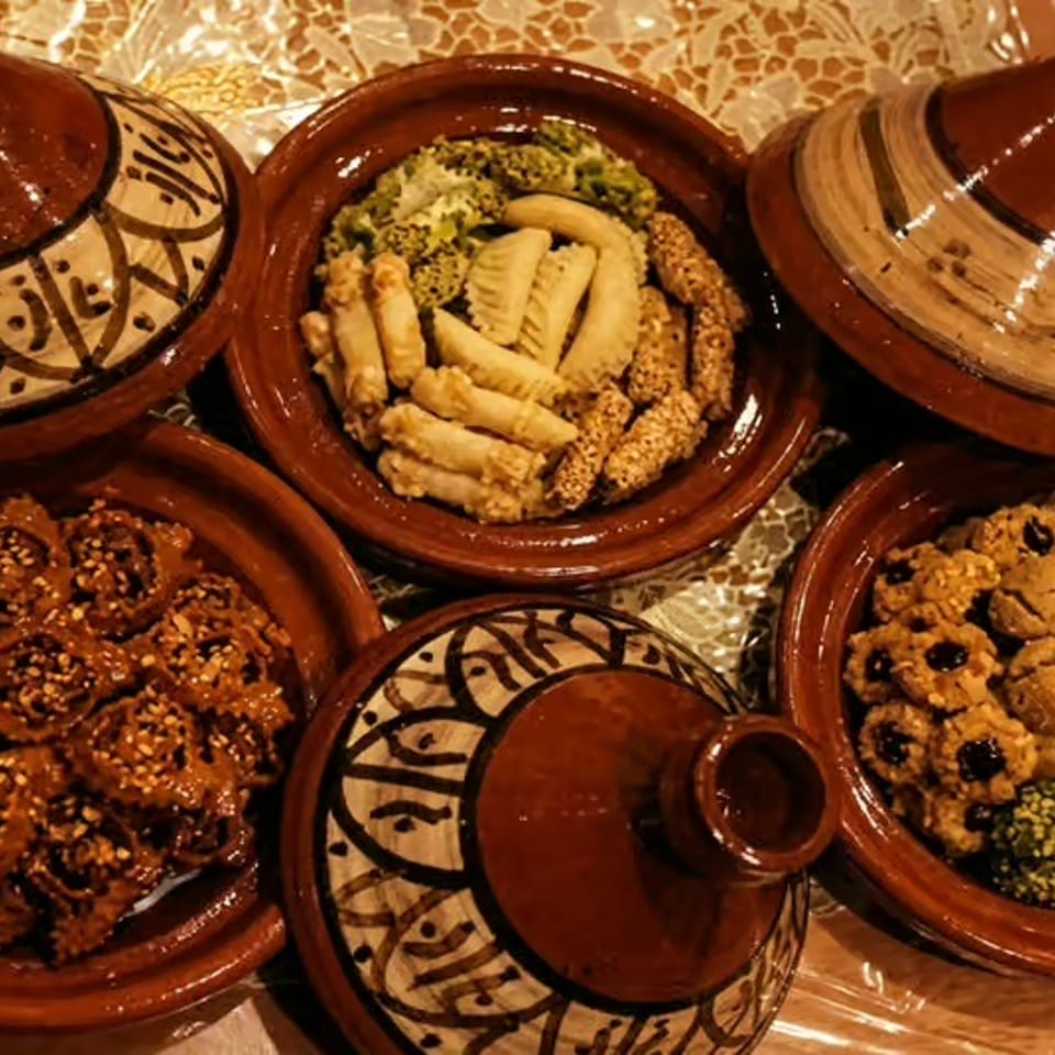 Moroccan tagine dishes and ceramic lids surround plates of assorted pastries and sesame-coated sweets on a patterned tablecloth.