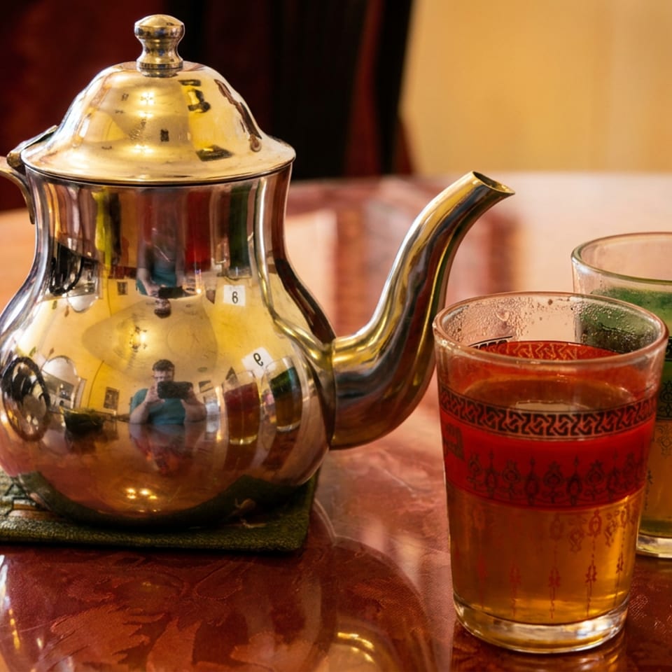 Ornate silver teapot beside two patterned glasses of amber and green tea on a polished wooden table.