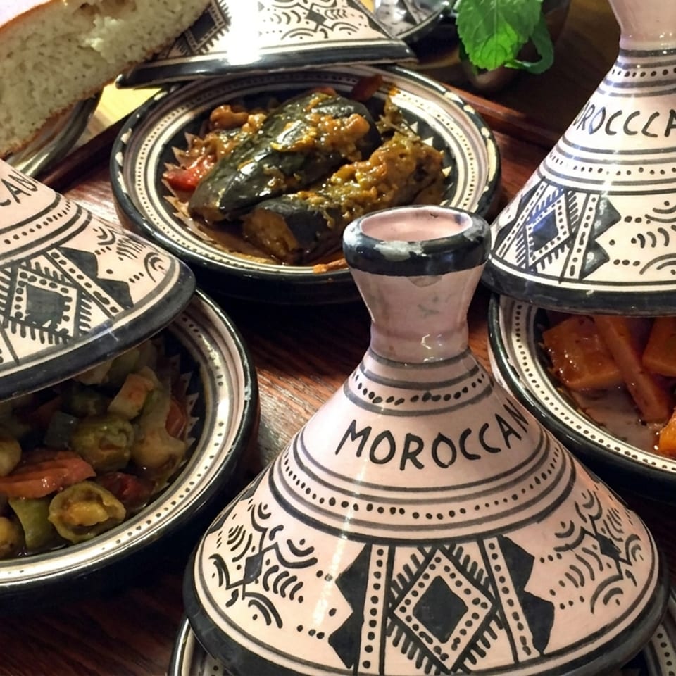 Four decorative Moroccan tagines surround dishes of colorful stewed vegetables and meat on a wooden table with bread.
