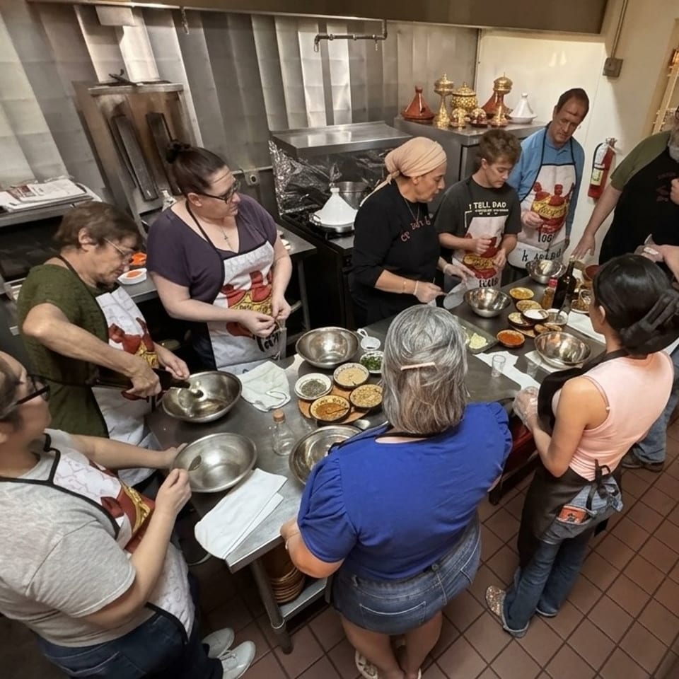 Group cooking class participants prepare food together around a restaurant kitchen table filled with bowls, spices, and ingredients.