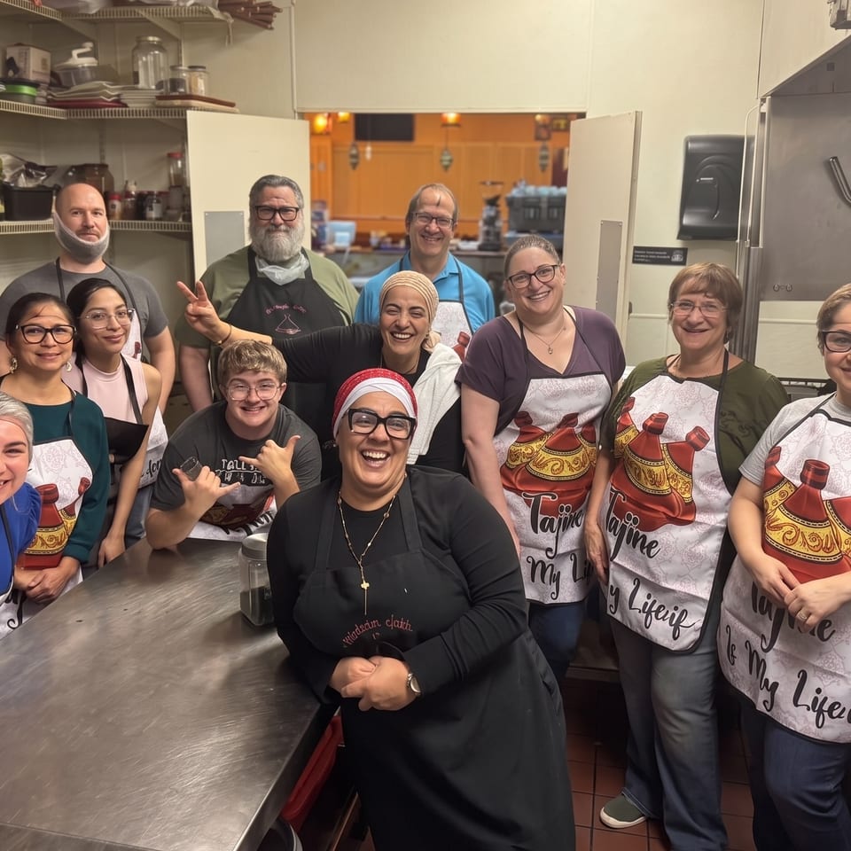 Smiling restaurant staff pose together in a commercial kitchen, wearing matching illustrated aprons around a stainless-steel prep table.