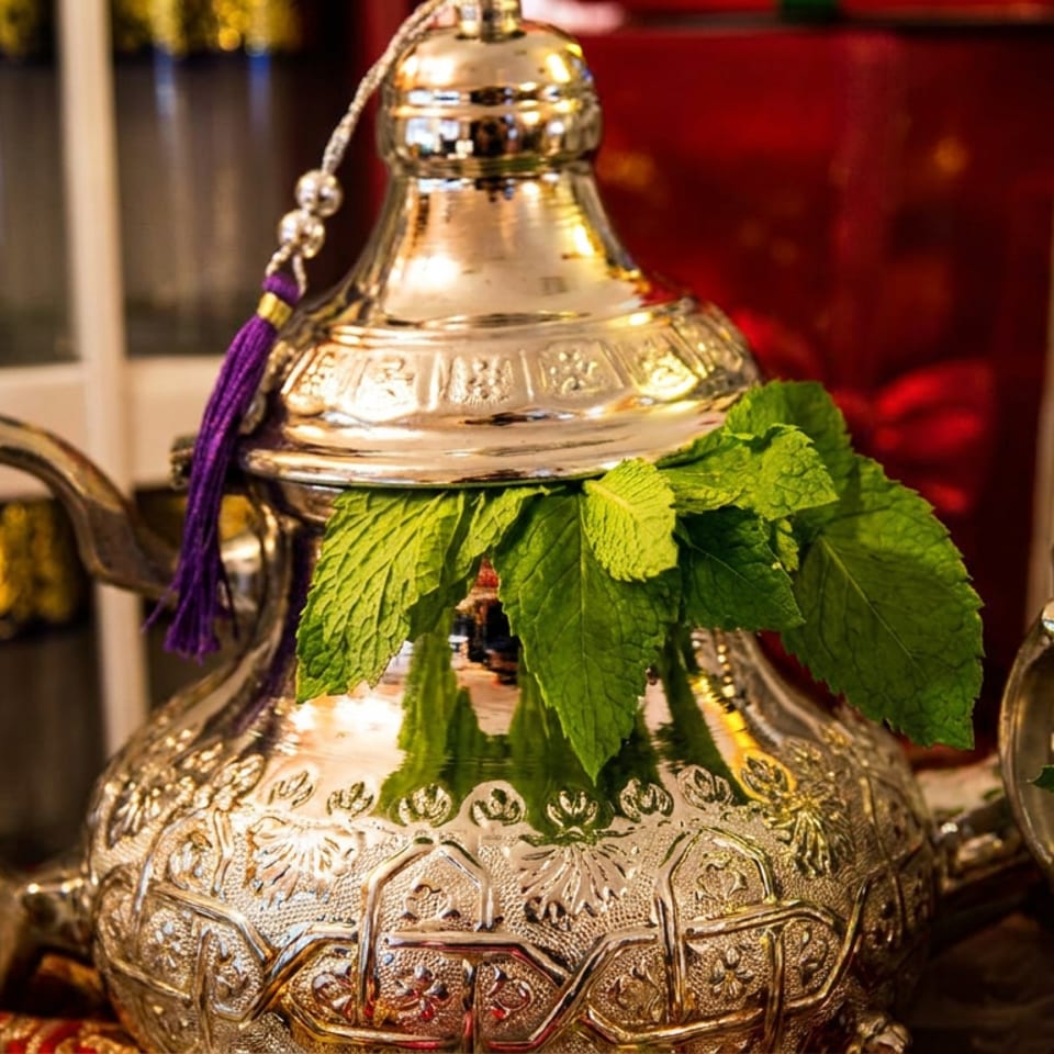 Ornate silver Moroccan teapot adorned with fresh mint leaves, surrounded by smaller teapots in a warmly decorated setting.