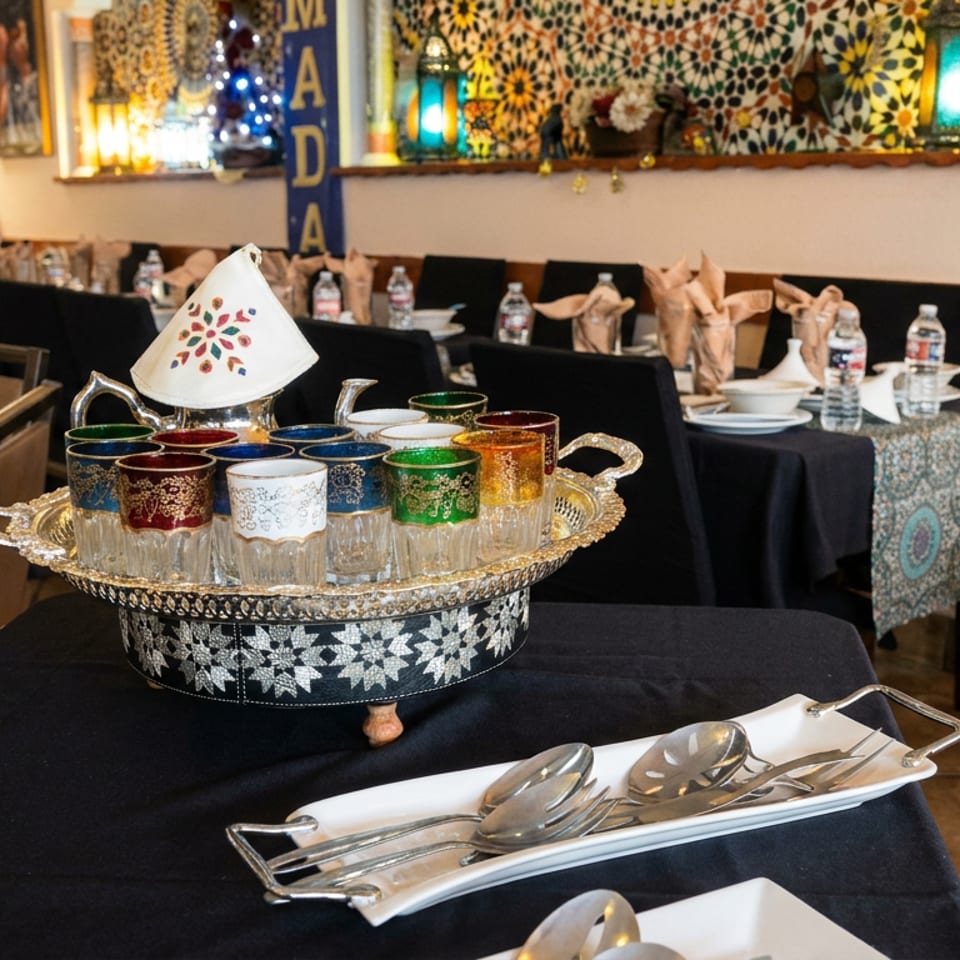 Ornate Moroccan tea glasses arranged on a silver tray in a warmly decorated restaurant with mosaic walls and set tables.