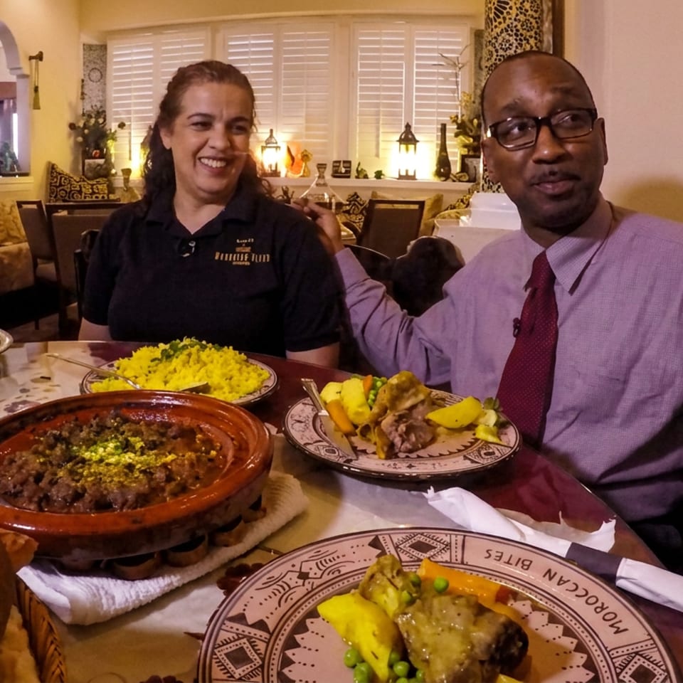 Two diners enjoy Moroccan dishes, including couscous, tagine, bread, and plated meat, in a warmly decorated restaurant.